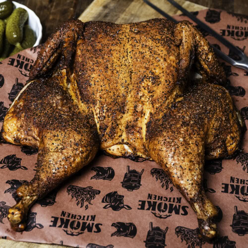 Smoked spatchcock chicken with crispy seasoned skin on butcher paper, served with pickles on a wooden board.