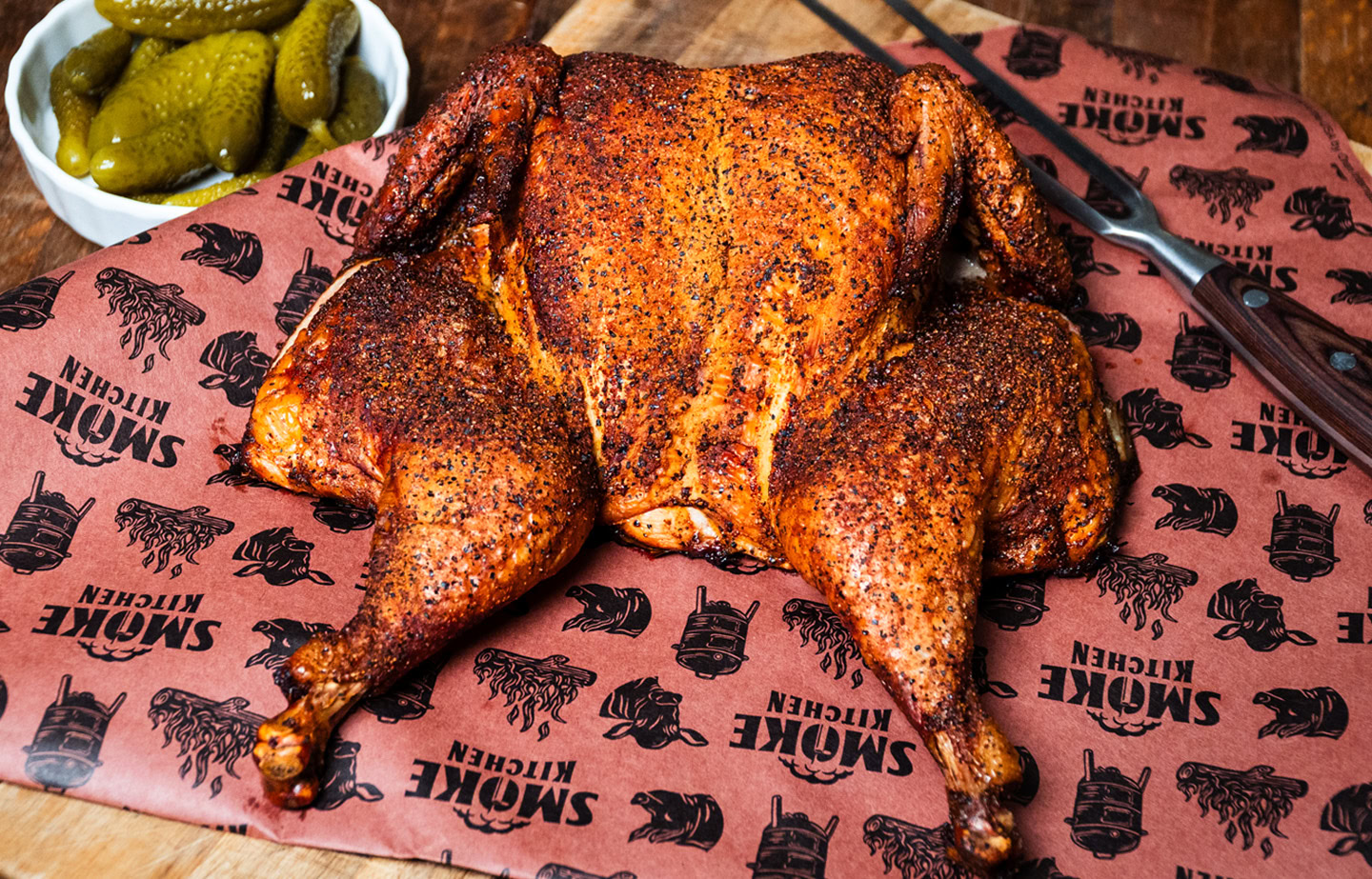 Smoked spatchcock chicken with crispy seasoned skin on butcher paper, served with pickles on a wooden board.
