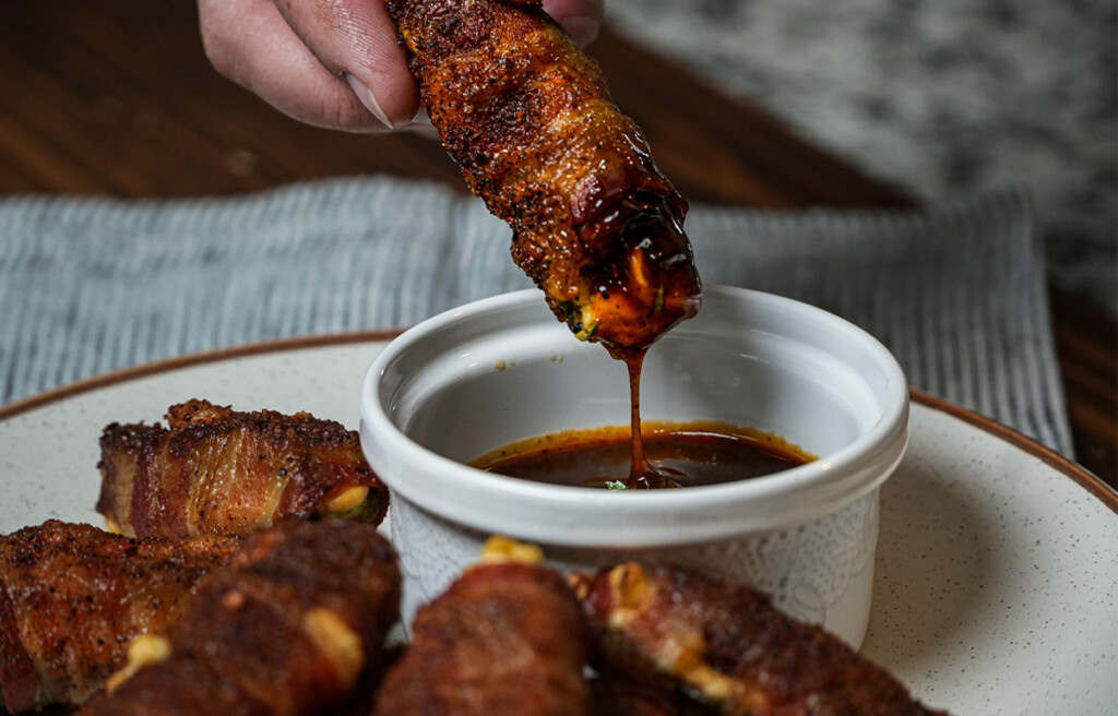 Hand dipping a bacon-wrapped jalapeño stuffed with cream cheese and sausage into barbecue sauce, with smoked Atomic Buffalo Turds arranged on a plate below.