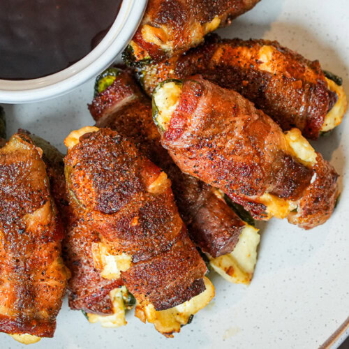 Bacon-wrapped jalapeño poppers stuffed with cheese, served on a white plate with barbecue sauce