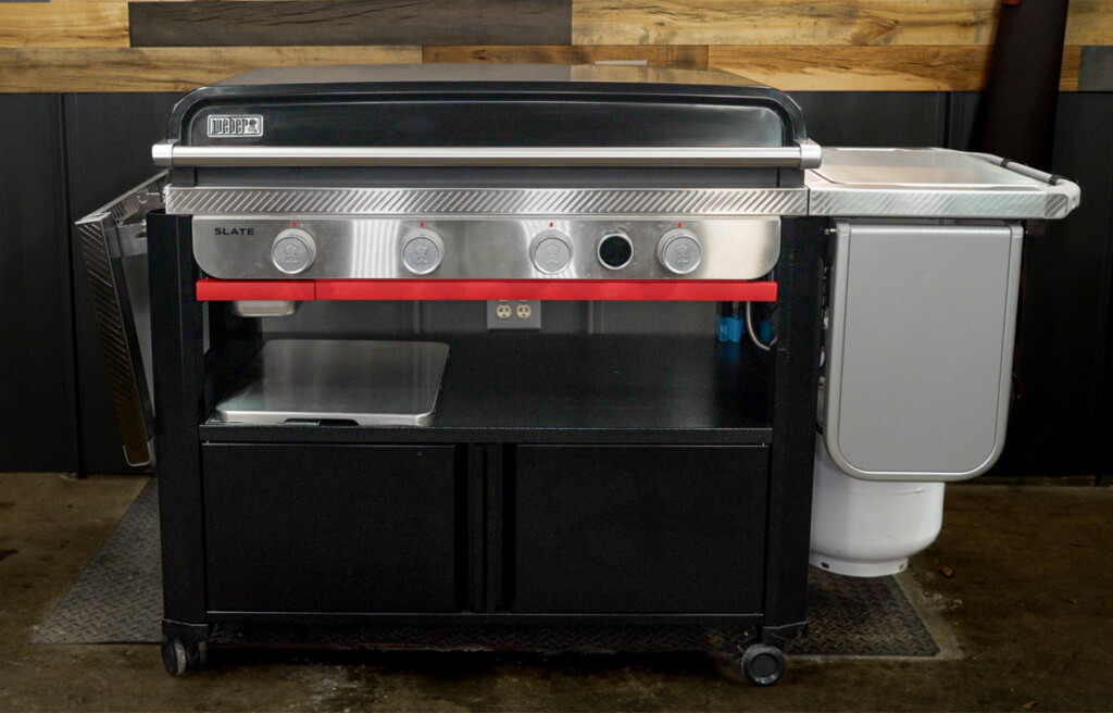 Front view of the Weber Slate outdoor gas griddle, featuring four control knobs, a solid lid, stainless steel trim, a side-mounted propane tank, fixed and foldable side shelves, and a lower cabinet for storage. The griddle is set up in a garage or workshop space with a wood-paneled wall in the background.