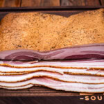 Slab of home-cured and smoked maple bacon with even marbling, shown sliced on a wooden cutting board with visible peppered crust and rich pink center.
