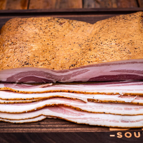 Slab of home-cured and smoked maple bacon with even marbling, shown sliced on a wooden cutting board with visible peppered crust and rich pink center.