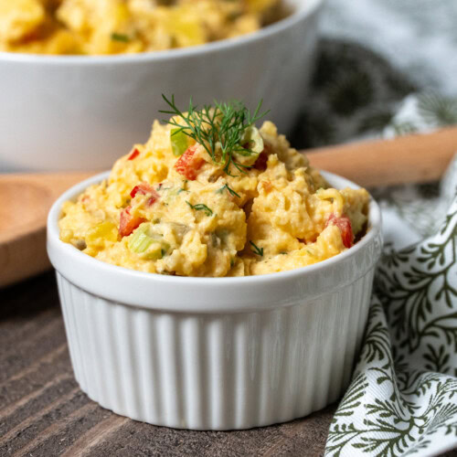 Creamy Southern potato salad served in a white ramekin, garnished with fresh dill and surrounded by a wooden spoon and patterned cloth napkin on a rustic wood surface.