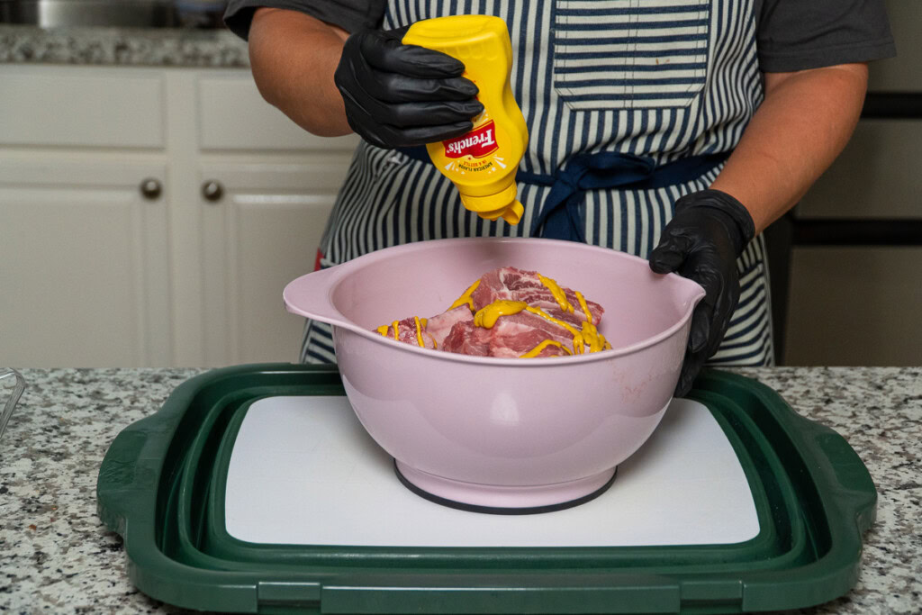a hand with black glove squeezing a bottle of yellow mustard onto a bowl of raw baby back ribs