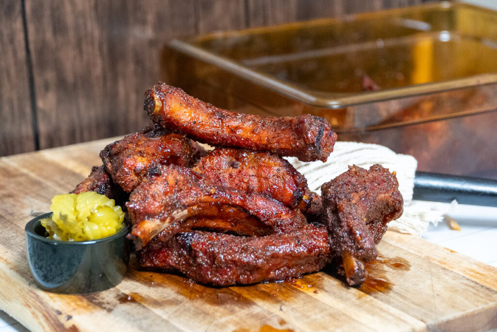 a stack of party ribs and a bowl of pickles on a wooden board
