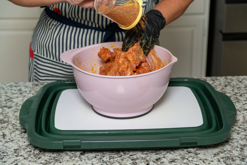 a black gloved hand holding a shaker of seasoning over a bowl of raw baby back ribs