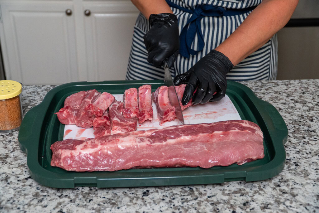a person with black gloves on, slicing raw baby back ribs into individual ribs