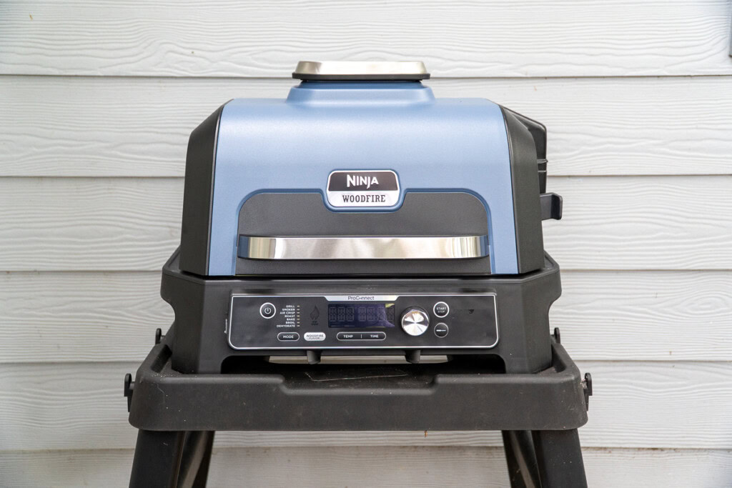 The Ninja Woodfire ProConnect XL electric grill in Coastal Blue, sitting on an outdoor stand. The front control panel features buttons for cooking modes, temperature, and time, along with a digital display and integrated dial.
