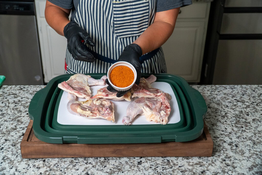 a person holding a pot of seasoning over a tray of raw chicken quarters