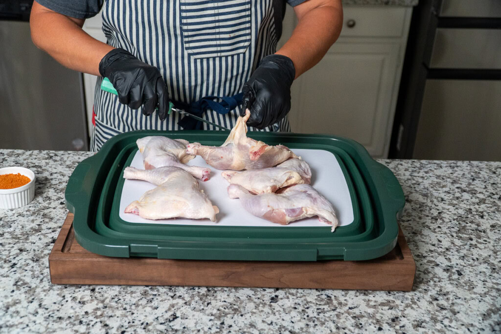 a person with black gloves trimming the excess skin off a raw chicken quarter