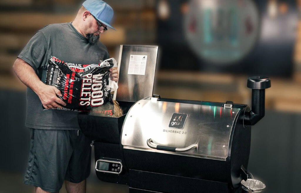 Man pouring a bag of wood pellets into the hopper of a Grilla Silverbac 2.0 pellet grill, with the stainless steel lid open and controller visible on the side.