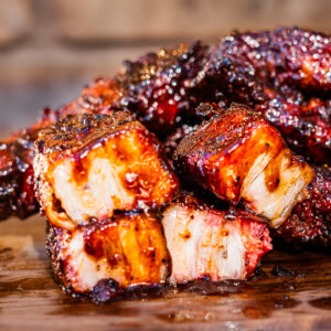 Close-up of smoked country style ribs on a wooden board with two broken pieces in the foreground revealing tender white and amber interior fat columns, surrounded by deeply glazed mahogany bark with juices pooling on the board.