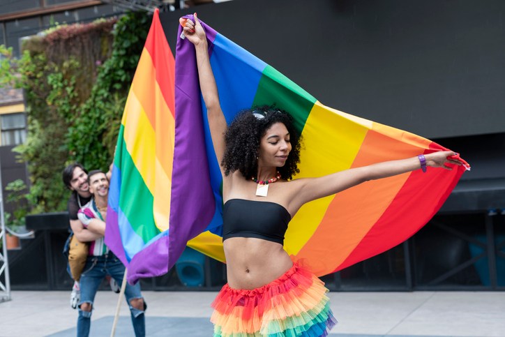 Portrait of a brunette Latina with curly hair enjoying her gay pride day dressed very colorful smiling at the camera while happily holding her gay pride flag