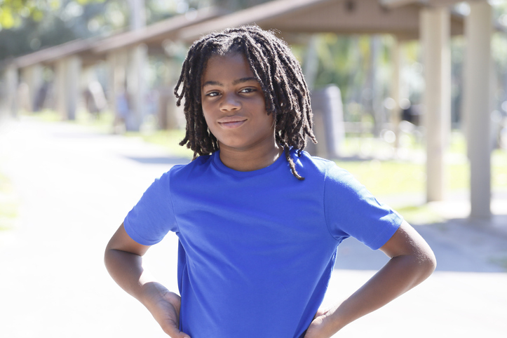 Portrait of a smiling young African American boy with locs, in an outdoor park, wearing a blue t-shirt.