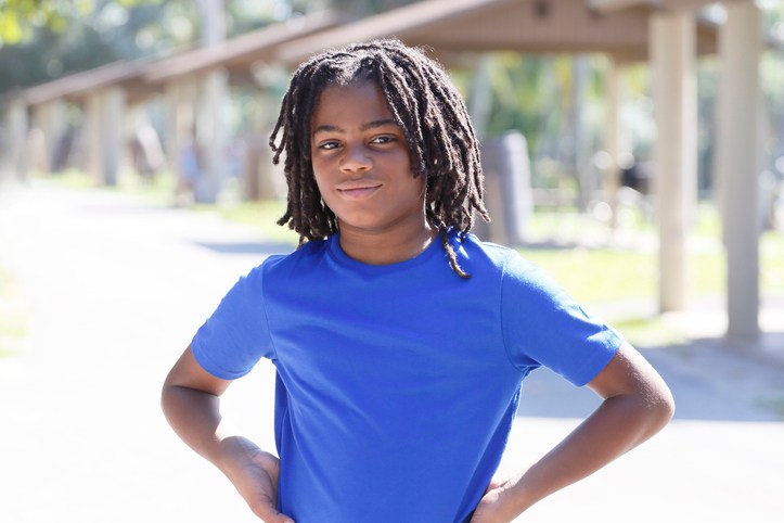 Portrait of a smiling young African American boy with locs, in an outdoor park, wearing a blue t-shirt.