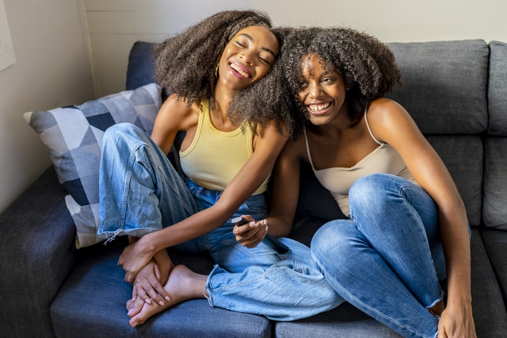 Young woman with remote control laughing by girlfriend in living room