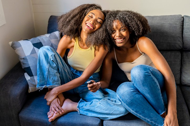 Young woman with remote control laughing by girlfriend in living room - holiday gift guide