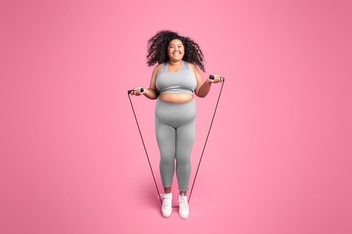 Happy african american chubby woman in sportswear jumping on jump rope, isolated on pink studio background, full length. survival mode