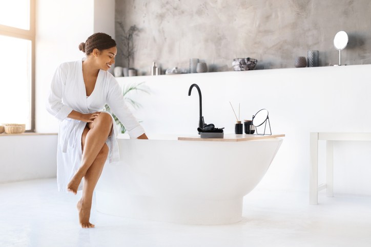 Graceful black woman in white bathrobe sitting by modern bathtub,