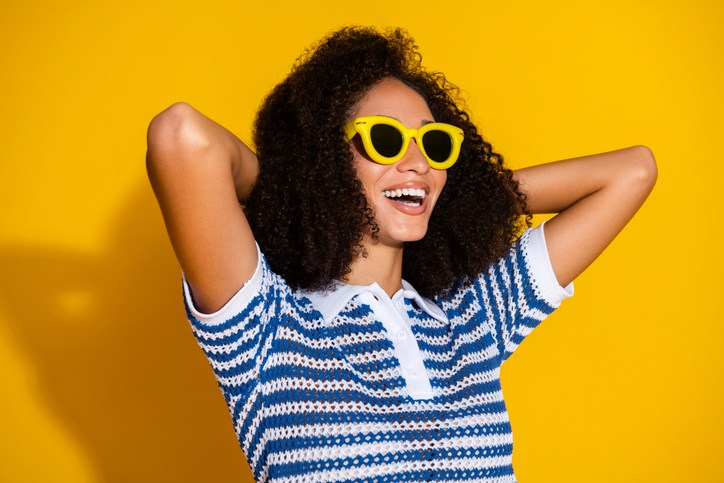 Stylish young woman with curly hair wearing yellow sunglasses and a striped polo shirt smiling