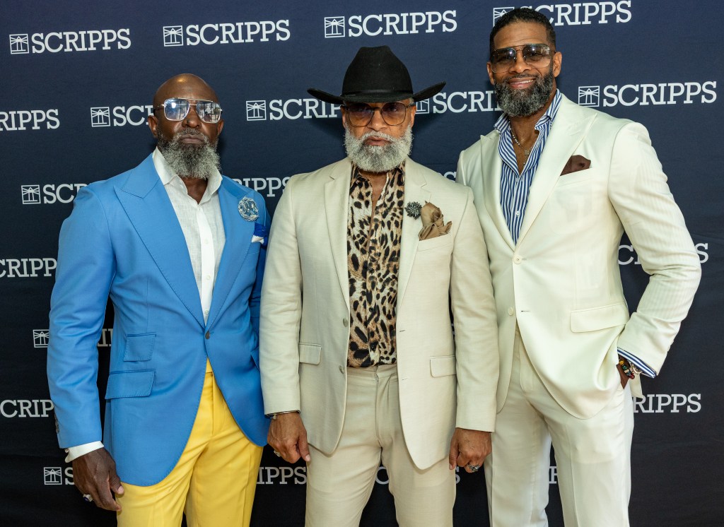 Three well-dressed men with beards and sunglasses posing in front of a Scripps backdrop.