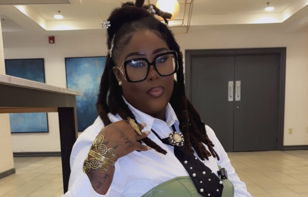 A person with long braided hair wearing glasses, a white shirt, and jewelry, standing in an indoor setting with artwork on the walls.