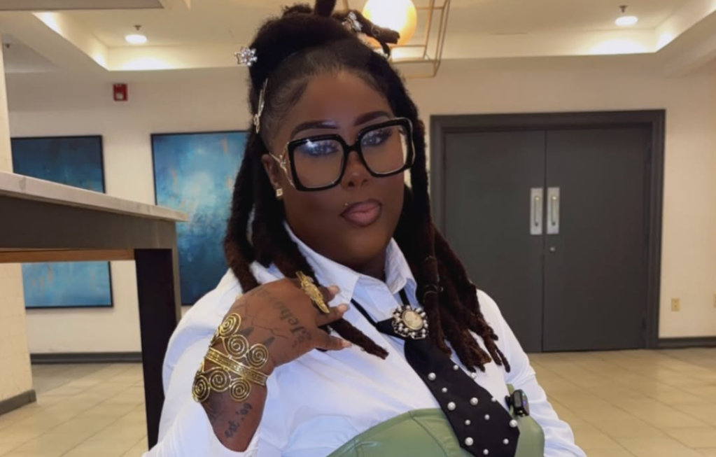 A person with long braided hair wearing glasses, a white shirt, and jewelry, standing in an indoor setting with artwork on the walls.