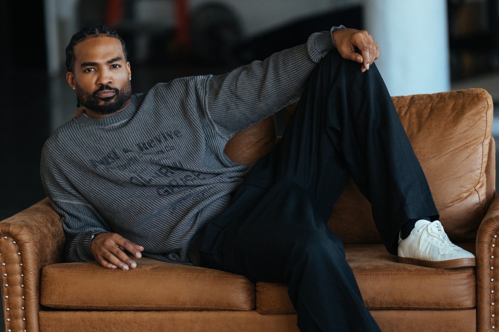 A man with a beard wearing a gray sweater sits on a brown leather couch, with his hands on his knees.