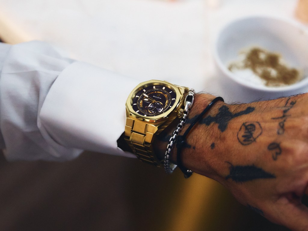A close-up view of a person's arm with a gold-colored watch and tattoos on the wrist.