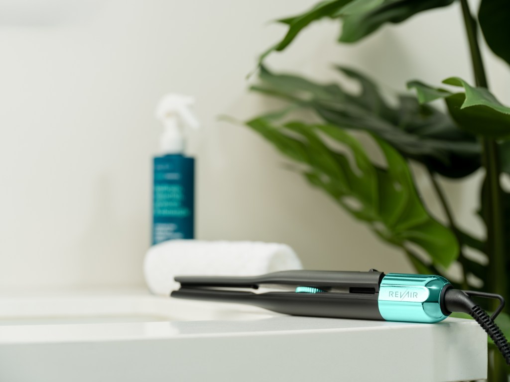 A green and black hair straightener with the brand name "Revair" on a white surface, surrounded by green leaves.