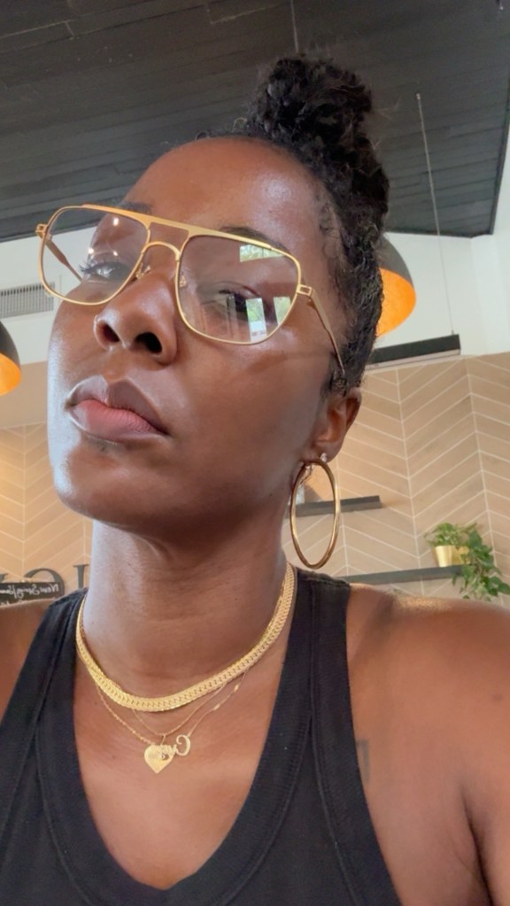 A close-up portrait of a young Black woman wearing large gold-framed glasses and a black top, with a serious expression on her face.