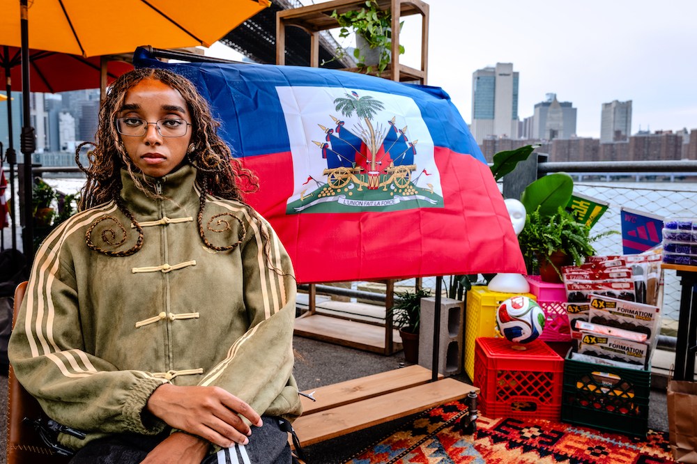 A person wearing a green jacket and glasses stands in front of a Haitian flag, surrounded by various items including a soccer ball and crates.