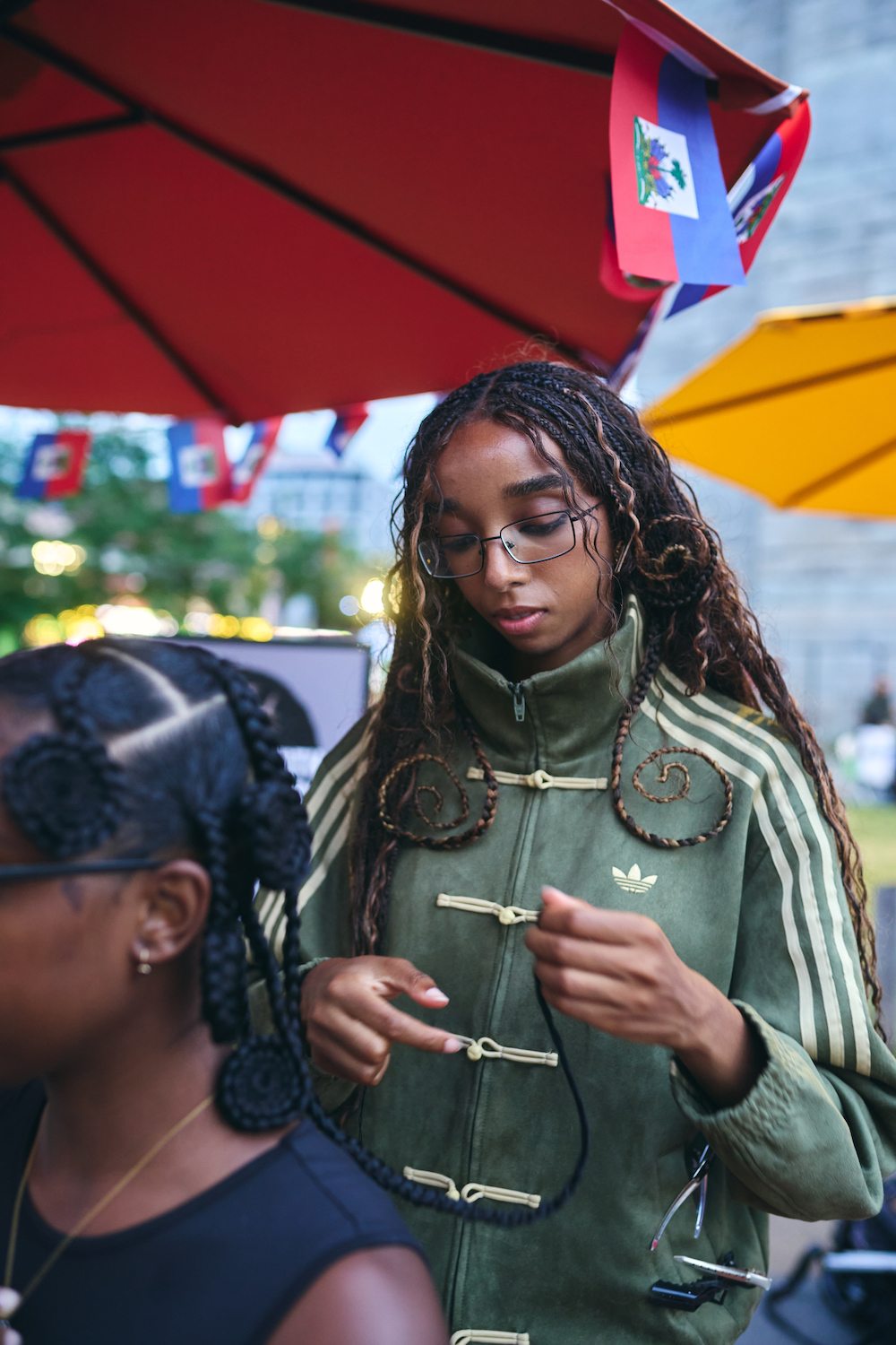 A person wearing an Adidas jacket with braided hair and glasses standing under a red and yellow umbrella in a crowded outdoor setting.