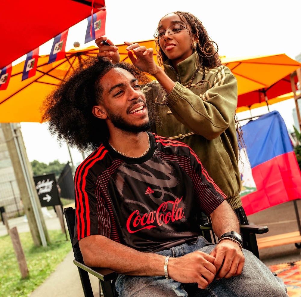 A person in a Coca-Cola jersey smiling while another person styles their hair in an outdoor setting with colorful umbrellas.