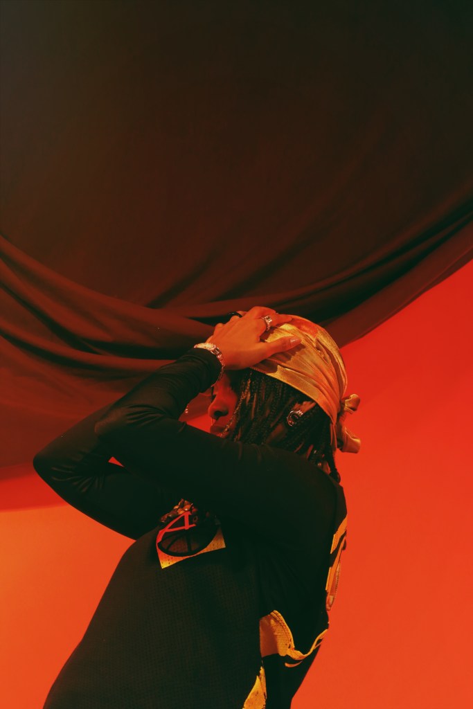 A person in a black outfit with a bright orange headpiece performing on a stage with dramatic red and green lighting.