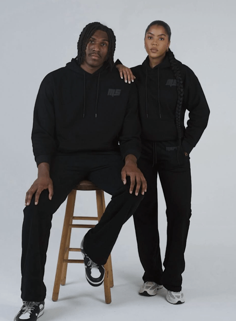 Two people wearing all-black clothing and posing together on a chair against a plain background.
