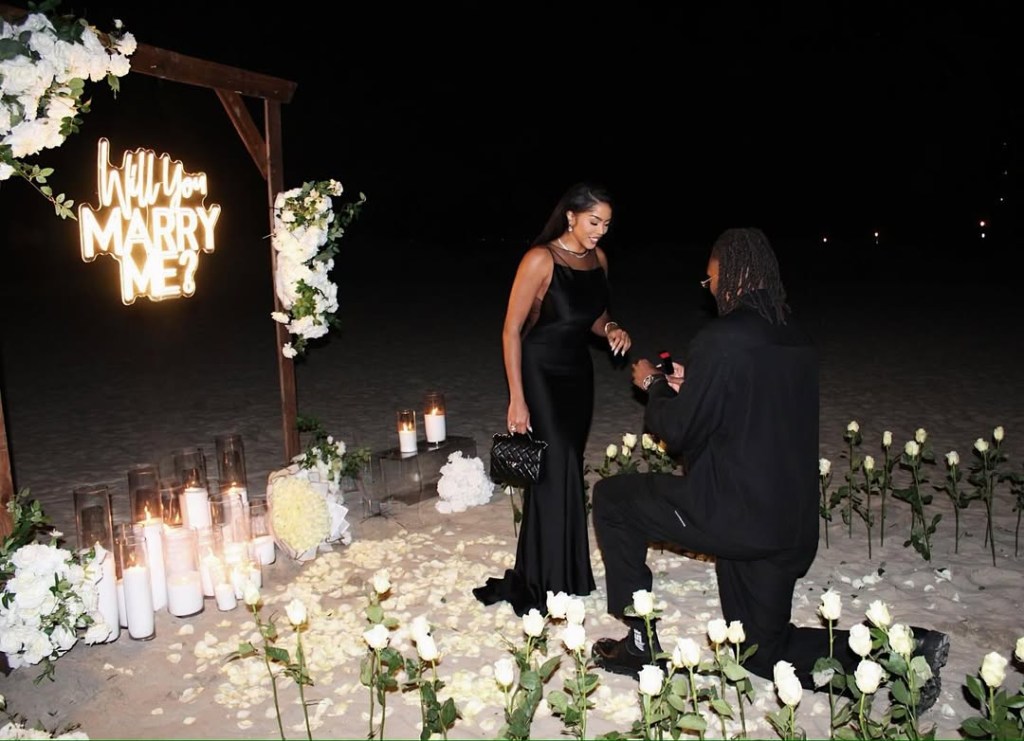 A couple standing in a romantic, flower-filled setting at night, with a neon "Will you marry me?" sign behind them.