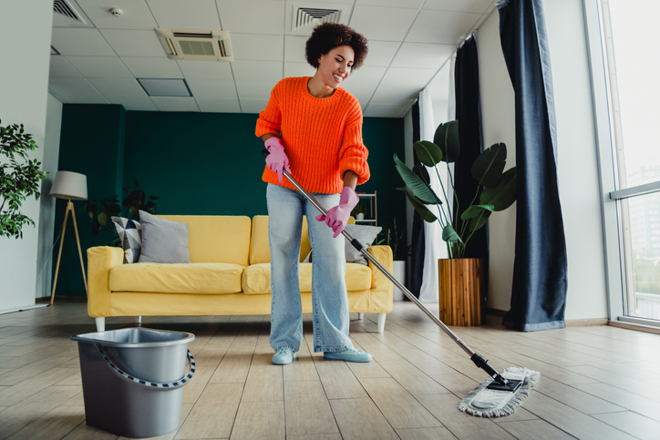 Happy woman mopping living room floor at home wearing bright sweater and pink gloves, domestic cleaning and household chore scene