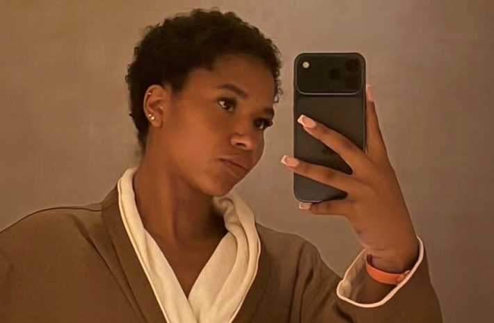A person wearing a beige robe holding a smartphone and looking at the camera.
