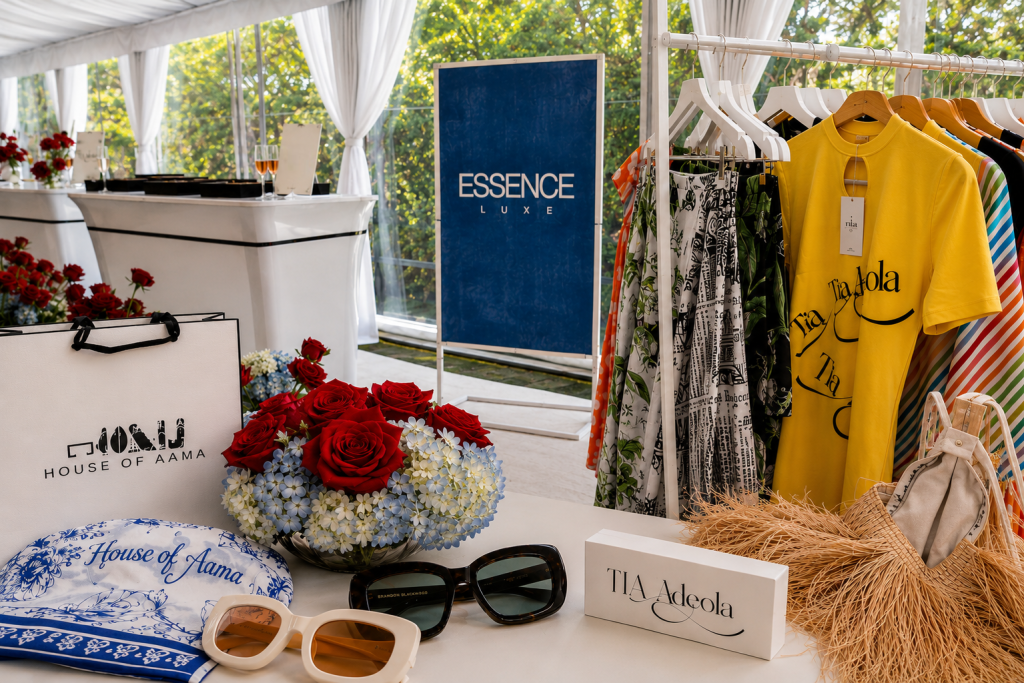 A display of clothing and accessories from the Essence Luxe brand, including a yellow t-shirt, floral dress, and sunglasses, set against a backdrop of flowers and a white tent.
