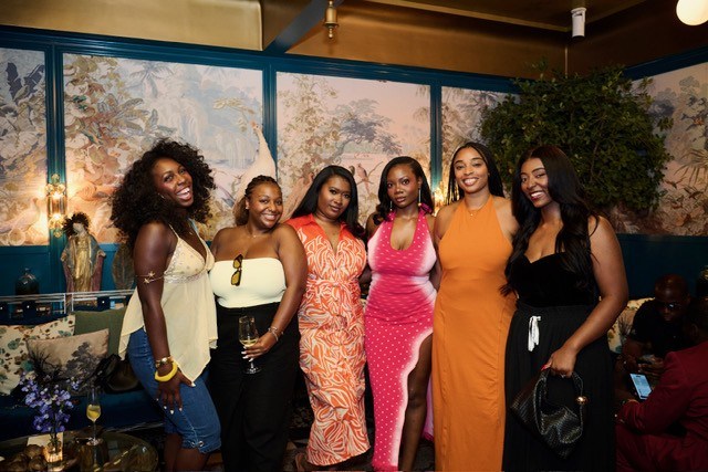 A group of six young women dressed in colorful, stylish outfits posing together in a festive setting with floral decor.