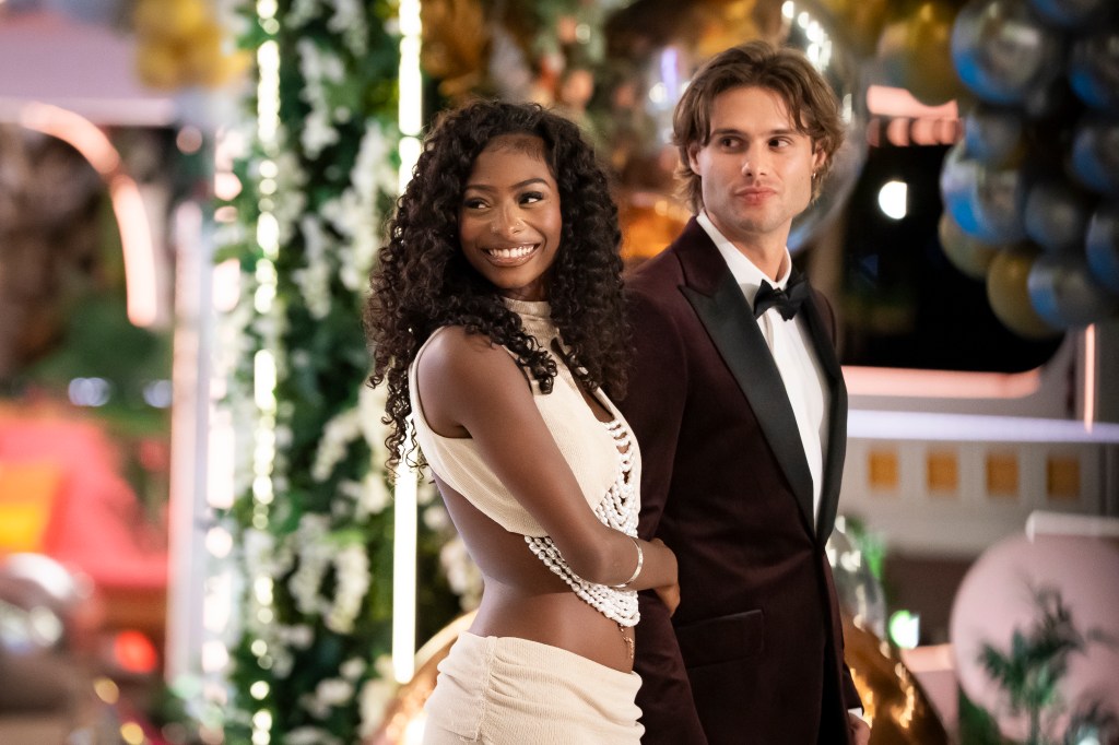 Two people, a smiling woman with curly hair and a man in a tuxedo, standing together in front of a decorated holiday setting.