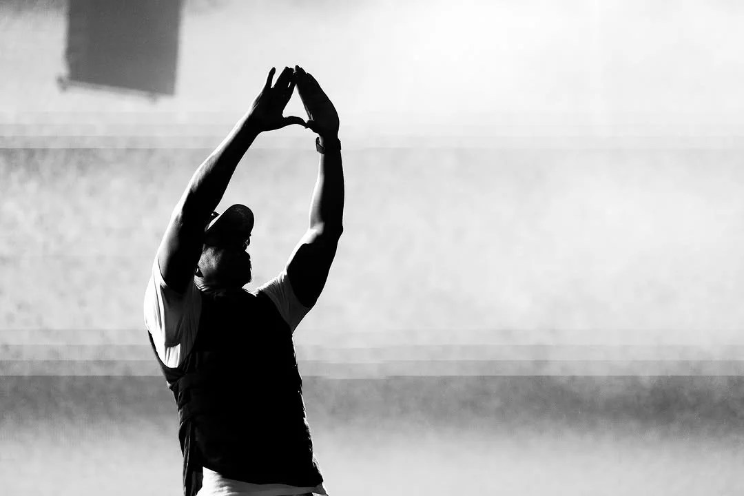 A person silhouetted against a hazy background, with their arms raised overhead in a stretching or reaching pose.