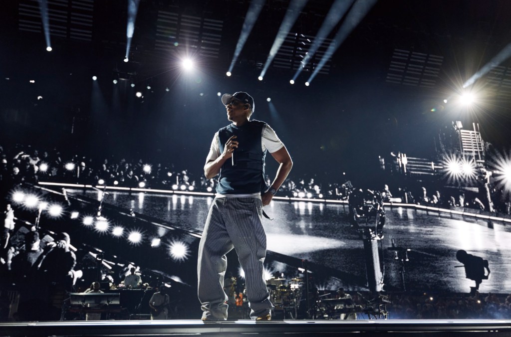 A performer on stage in a dimly lit concert venue, surrounded by a crowd of people and bright stage lights.