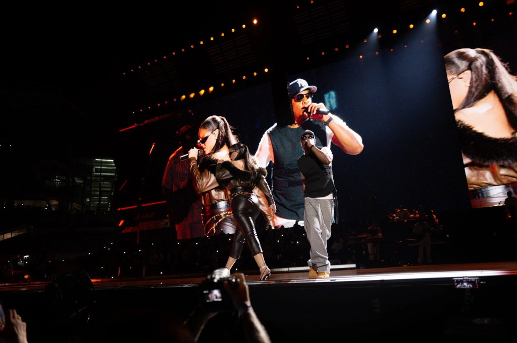 Two performers on stage, one wearing a leather jacket and sunglasses, the other in a red outfit, singing and dancing under bright stage lights.