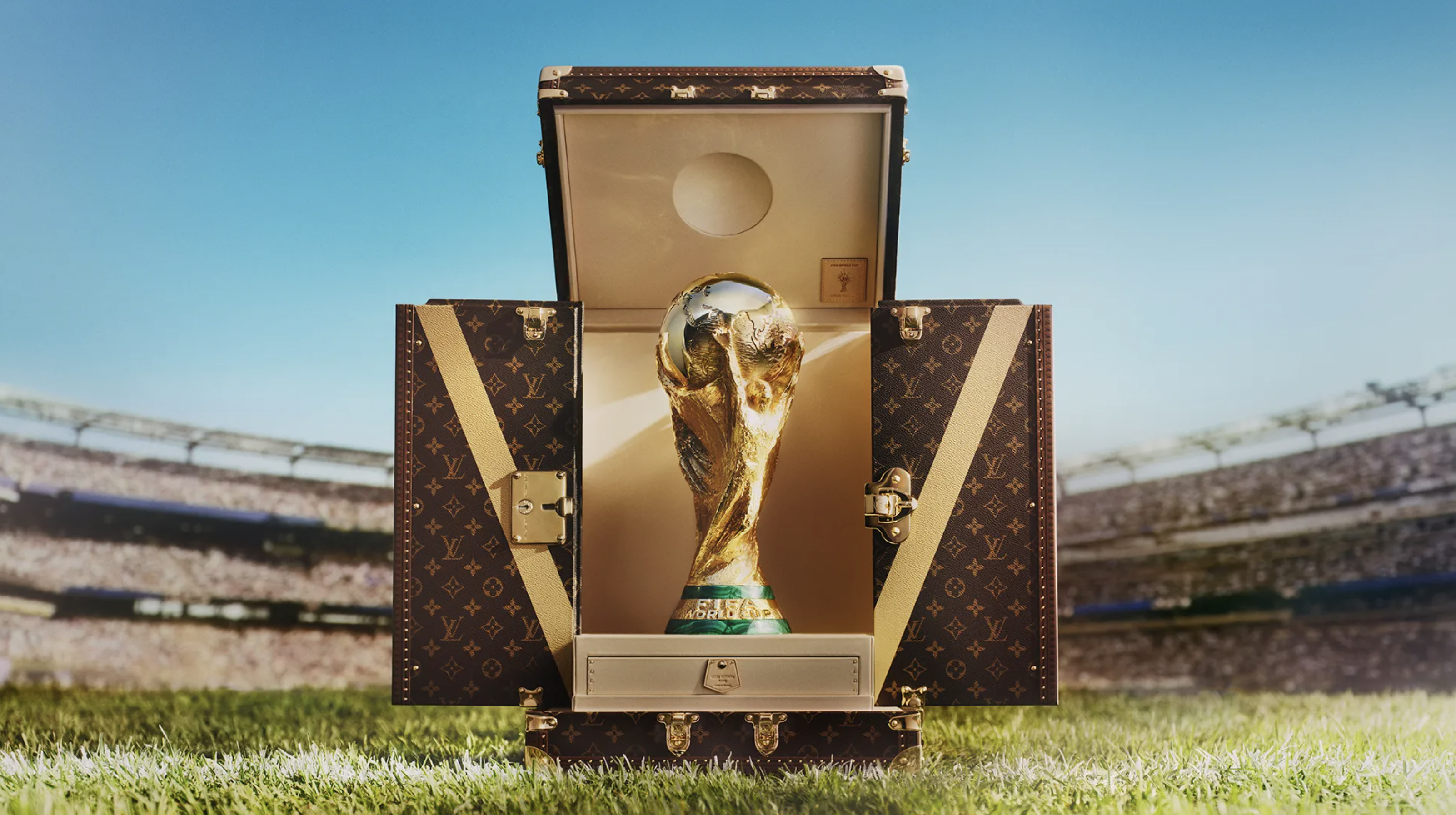 A golden trophy representing the FIFA World Cup is displayed in a Louis Vuitton-branded trunk on a grassy field.