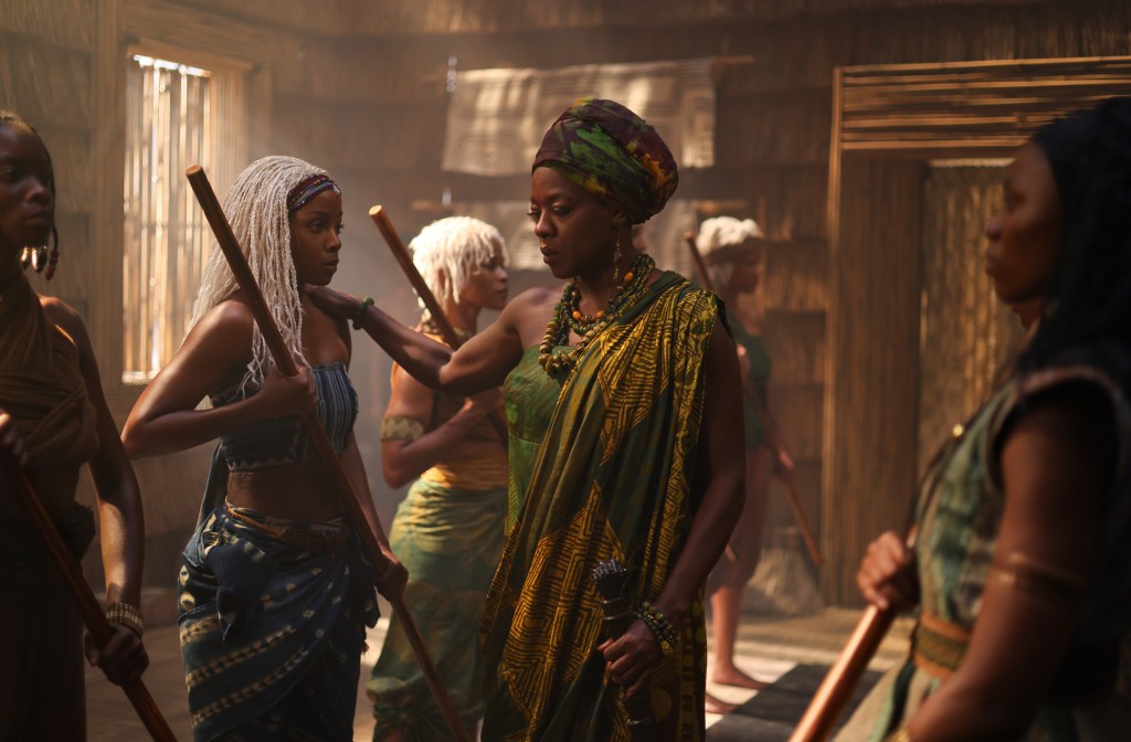 A group of people, including two women in colorful traditional African attire, stand in a dimly lit room. One woman holds a staff.