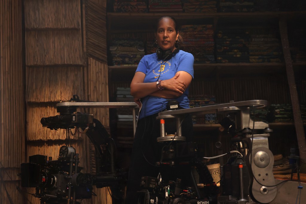 A woman in a blue t-shirt stands with her arms crossed in a room filled with film equipment, including cameras and lighting gear.