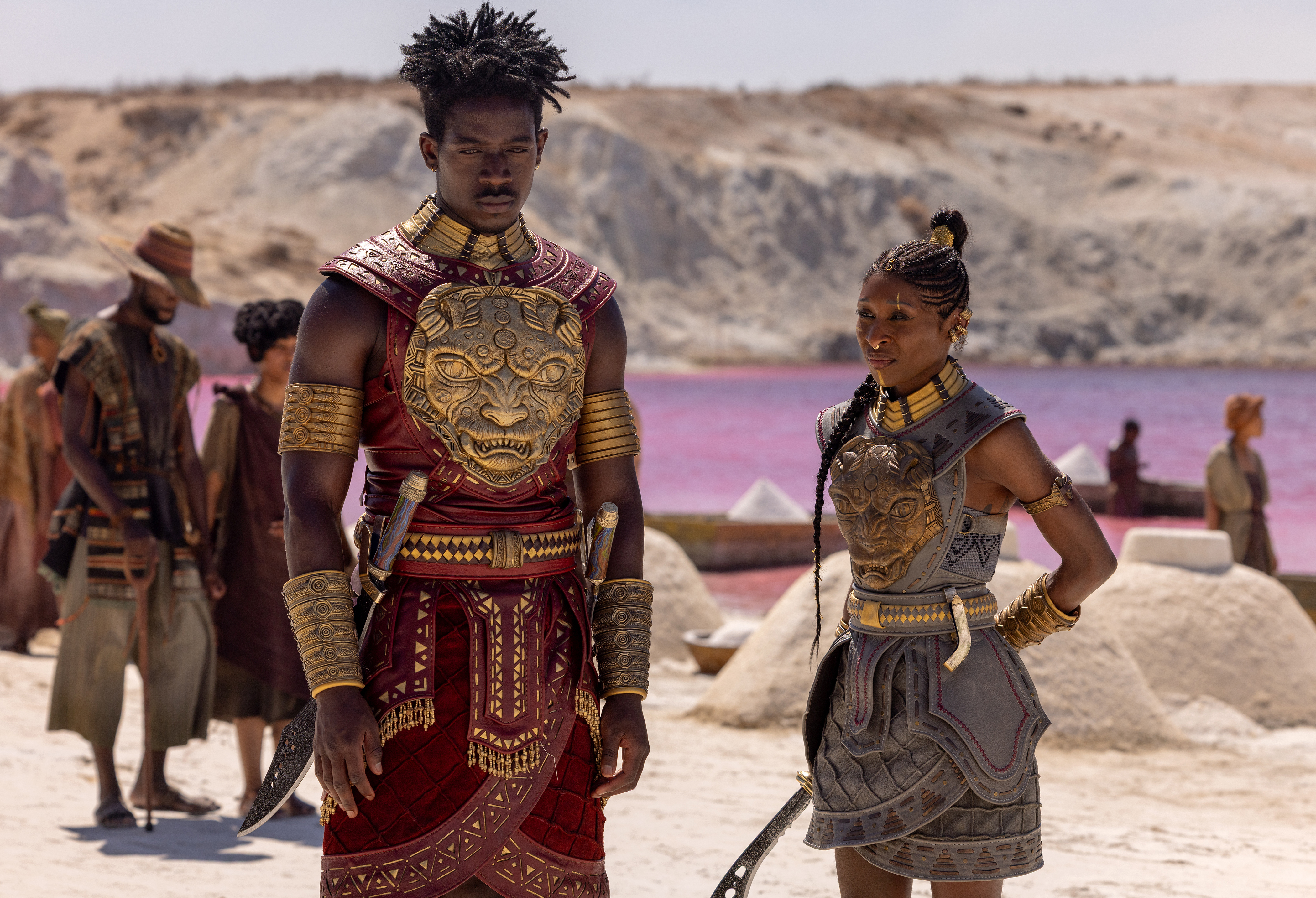 Two people in elaborate tribal costumes stand in a desert landscape with a pink lake in the background.